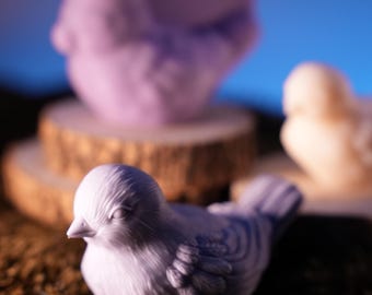 May include: A close-up of three bird figurines in a studio setting. The foreground features a light purple bird on a bed of brown moss. In the background, a light purple bird is perched on a wooden disc, and a beige bird is in the background.