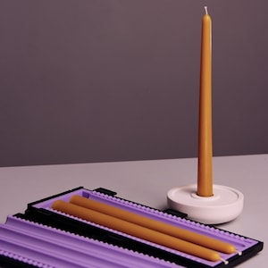 Op de afbeelding: Een lange, slanke, gele kaars in een witte keramische houder. Twee extra gele kaarsen liggen in een zwarte en paarse opbergdoos. De achtergrond is effen paars.