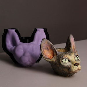 May include: A lavender silicone mold shaped like a Sphynx cat head positioned behind a finished, hand-painted Sphynx cat head sculpture with green eyes, mottled brown and tan fur textures, and pink-tinted ears, both featuring pointed ears.