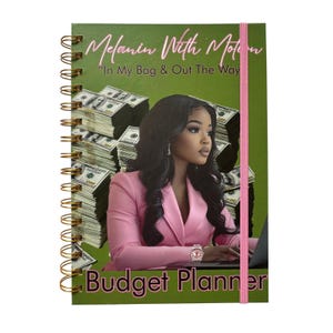 May include: A spiral-bound budget planner with a green cover featuring a woman in a pink blazer, stacks of money, and the text "Melanin With Motion" and "Budget Planner". A pink elastic band secures the planner.
