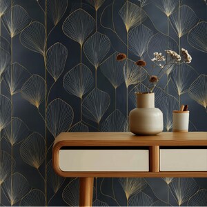 Könnte beinhalten: Dunkelblaue Tapete mit goldenem Blattmuster. Ein Holztisch mit zwei Schubladen trägt eine beige Vase mit getrockneten Blumen und einen kleinen weißen Becher mit Bleistiften. Das Blattmuster wiederholt sich im Bild.