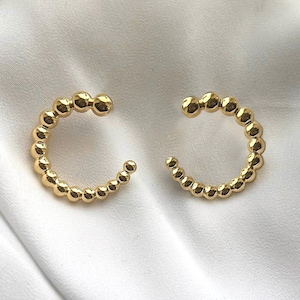 May include: A pair of gold-colored hoop earrings. Each earring is a half-circle design, crafted from small, evenly spaced beads. The earrings are displayed on a white, textured surface, creating a simple and elegant presentation. The earrings are a jewelry item.