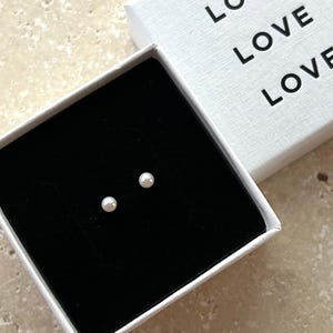May include: A pair of small, round pearl earrings displayed in an open white box with the word "LOVE" printed three times. The earrings sit on a black velvet interior, and the box is on a light-colored surface.