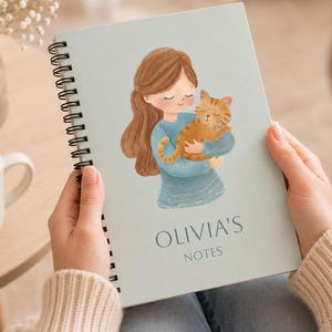 Può includere: Un quaderno a spirale azzurro con un'illustrazione ad acquerello di una persona con lunghi capelli castani che tiene in braccio un gatto tigrato arancione. Il testo "OLIVIA'S NOTES" è stampato in blu scuro sotto l'illustrazione sulla copertina.