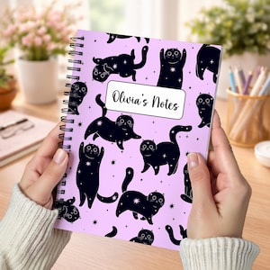 May include: A spiral-bound notebook with a light purple cover featuring a repeating pattern of black cats with white star accents. A white rectangular label in the center contains the black text Olivia's Notes in a cursive script font.