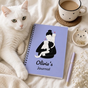 May include: A light purple spiral-bound journal featuring a graphic illustration of a person holding two white cats with small red hearts above them. The text on the cover reads Olivia's Journal in black lettering, positioned below the illustration.