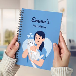 以下が含まれることがあります： 「Emma's Vet Notes」の文字が入った、水色のスパイラルノート。表紙には、白い猫を抱きかかえる人物のイラストと、3つの小さなハートが描かれています。ノートは両手で持たれています。
