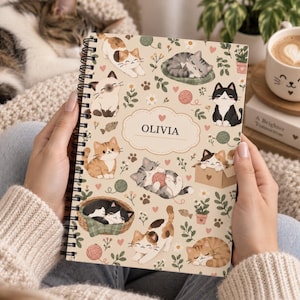 May include: A spiral-bound notebook with a beige cover featuring a repeating pattern of various cats, yarn balls, and small plants. The center of the cover displays a cloud-shaped label with the name OLIVIA printed in black capital letters.