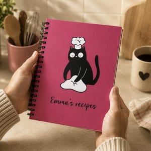 May include: A spiral-bound notebook with a magenta cover featuring a black cat wearing a white chef hat and kneading dough. The text "Emma's recipes" is printed in black script below the cat illustration. The notebook has a black metal coil.