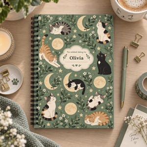 Peut inclure: Un carnet à spirales avec une couverture vert sauge ornée d'un motif de chats, de lunes, d'étoiles et de fleurs blanches. Une étiquette décorative centrale porte le texte "This notebook belongs to: Olivia" en lettres noires.