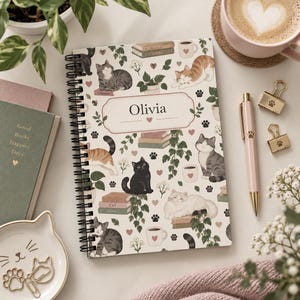 May include: A spiral-bound notebook featuring a pattern of cats, stacks of books, coffee mugs, and small hearts. The centre of the cover has a rectangular label with the name Olivia printed in black text. The background is off-white.