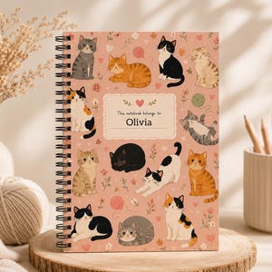 May include: A spiral-bound notebook with a dusty pink cover featuring a pattern of various cats, small flowers, and yarn balls. A central rectangular label with a heart icon contains the printed text: This notebook belongs to: Olivia.