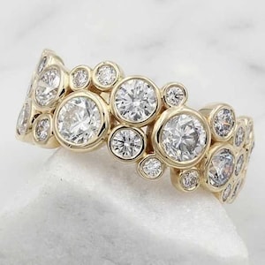 May include: A gold ring featuring multiple round, clear gemstones of varying sizes. The gemstones are set in individual bezels, creating a cluster design. The ring is displayed on a white surface, showcasing its sparkle and intricate details.