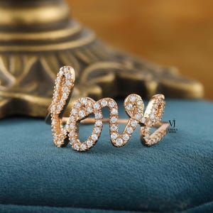 May include: Rose gold ring spelling out the word "love" in cursive script. The ring is encrusted with small, clear gemstones. The ring is displayed on a teal surface, with a blurred gold-coloured object in the background.