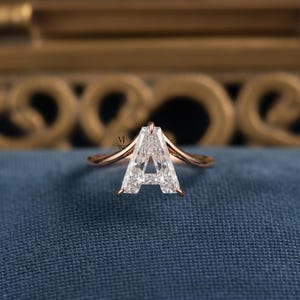 Puede incluir: Un anillo de oro rosa con una gema transparente en forma de letra "A". El anillo tiene una banda delicada que se curva hacia arriba para encontrarse con la gema. El anillo está colocado sobre una superficie azul con un fondo dorado borroso.