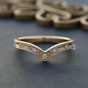 May include: A gold-colored ring with a V-shaped design. The ring features a delicate leaf pattern and small, sparkling gemstones. The ring is set against a dark blue background, highlighting its intricate details.