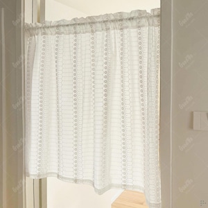 May include: A white lace curtain with a delicate floral pattern hangs in a doorway. The curtain is gathered at the top and has a scalloped edge. The light filters through the intricate design, creating a soft, airy feel.