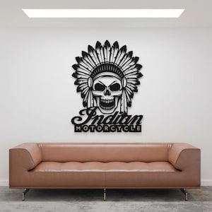 Może przedstawiać: Czarna metalowa grafika ścienna przedstawiająca czaszkę w indiańskim nakryciu głowy. Poniżej znajduje się napis "Indian Motorcycle". Grafika jest zamontowana na białej ścianie nad brązową skórzaną sofą.
