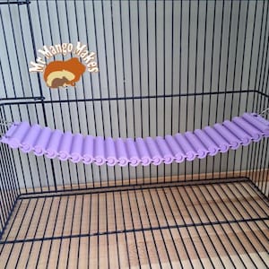 May include: A purple hanging hammock for small animals, made of a series of connected tubes, is suspended inside a wire cage.