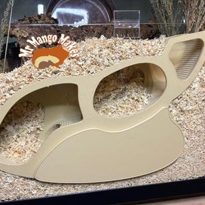May include: A beige, multi-chambered hamster hideout with a curved design. The hideout features multiple openings and a textured surface, providing a safe and engaging space for small pets. The Mr. Mango Makes logo is visible.