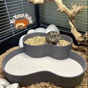 May include: A grey hamster in a multi-level habitat with a sand bath and a food bowl filled with wood shavings. The cage includes a decorative branch and the logo "Mr. Mango Makes".