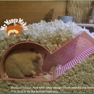 May include: Pink hamster hide with a daisy design. The hide is made of plastic and has a ramp leading up to the burrow. The burrow is large enough for a hamster to fit inside. The hide is sitting on a bed of white shredded paper.