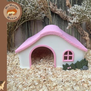 May include: A small white house-shaped pet hideout with a textured pink roof, a large arched entrance, and a small window with a pink frame. The structure features decorative green foliage and small pink flowers at the base of the front wall.