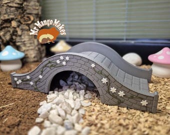 May include: A gray, arched bridge with a stone-like texture, adorned with white flower accents and climbing vines. The bridge is designed for small pets and is surrounded by decorative mushrooms and small rocks. The bridge has a curved ramp.