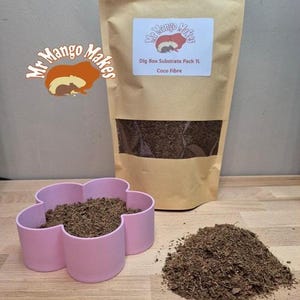May include: A brown paper bag with a clear window, a pink flower-shaped container, and a pile of brown coco fibre substrate. The bag has a label that reads "Dig Box Substrate Pack 1L Coco Fibre" and a logo that says "Mr Mango Makes".