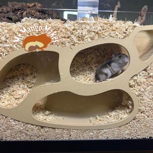 Peut inclure: Un habitat beige pour hamster avec plusieurs ouvertures et une rampe. Un petit hamster est à l'intérieur d'une des ouvertures. L'habitat porte le texte "Mr. Mango Makes" et un logo de hamster de dessin animé.