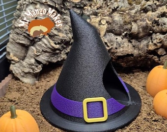 May include: A black witch hat with a purple band and yellow buckle, designed for a small pet. The hat is surrounded by orange pumpkins and a textured background, creating a Halloween theme.