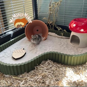 May include: A hamster habitat with a green scalloped border, filled with white sand. It includes a terracotta pot, a red mushroom house, and a wooden acorn. A small grey hamster is in the habitat.