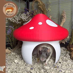 May include: A red and white mushroom-shaped pet house with a small rodent inside. The mushroom has a red top with white spots and a white base. The pet house is in a cage with bedding and dried plants.