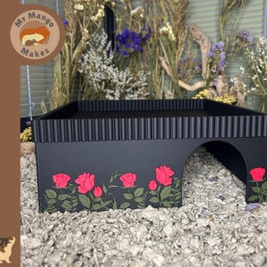 May include: A matte black rectangular animal hideout featuring a corrugated top edge and a side archway entrance. The front panel is decorated with a detailed graphic of vibrant red roses and green stems, leaves, and foliage across the base.