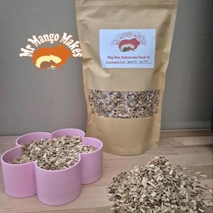May include: A brown paper bag labelled "Mr Mango Makes" with a clear window showing wood chips. A pink flower-shaped container and a pile of wood chips are in front of the bag. The text on the bag says "Dig Box Substrate Pack 1L".
