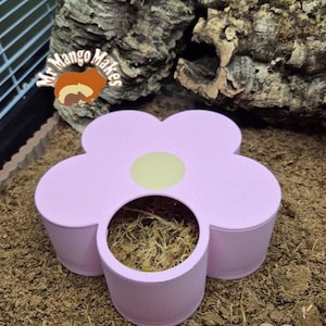 May include: A pink, flower-shaped pet habitat accessory with a central opening. The accessory has a light green circle in the centre and is filled with moss. The background includes a cage, bark, and substrate.