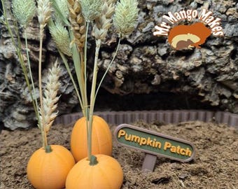 May include: Three orange pumpkins with green stems are arranged on a bed of brown soil, with wheat stalks and green foliage. A sign that reads "Pumpkin Patch" is in the foreground. The background features a textured, brown surface.