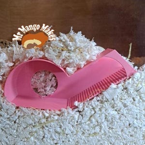 May include: Pink plastic hamster tunnel with a textured surface and a round opening. The tunnel is partially buried in white shredded paper.