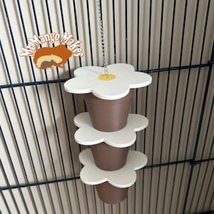 May include: A hanging pet toy featuring three brown flower pot-shaped containers stacked with white flower-shaped platforms. The toy is suspended by a silver chain and is designed to be hung inside a cage.