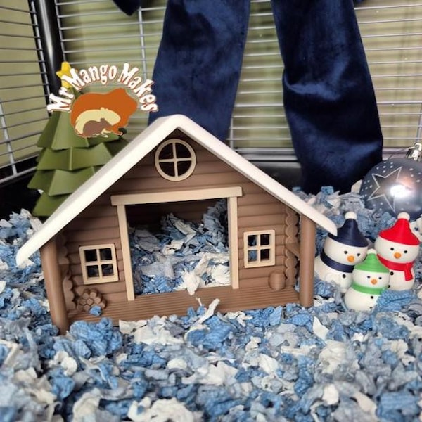 May include: A brown miniature cabin with a white roof, small windows, and a round window. The cabin sits on a bed of blue and white paper bedding, with small snowman figurines nearby. A sign that says "Mr. Mango Makes" is also visible.