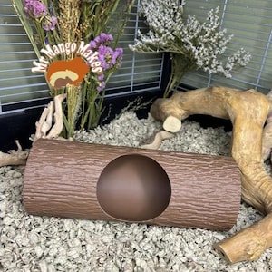 Peut inclure: Un abri pour animaux de compagnie en forme de bûche marron avec une grande ouverture circulaire. L'abri a une surface texturée ressemblant à de l'écorce d'arbre. L'image comprend également des fleurs séchées et une cage en arrière-plan.