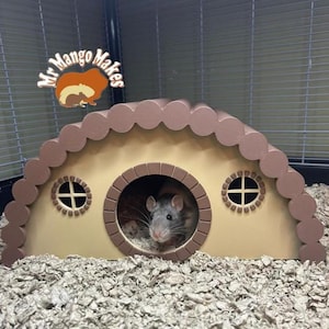 May include: A decorative pet house in tan and brown, featuring a round doorway and two small round windows. A grey rat is visible inside the doorway. The house is in a cage with paper bedding. The logo "Mr Mango Makers" is in the top left corner.