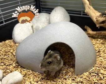 May include: A small, gray hamster peeks out from a gray, stone-like hideaway in a cage filled with wood shavings. The cage also contains white, egg-shaped stones and a piece of driftwood. The logo "Mr Mango Makes" is visible in the upper left corner.