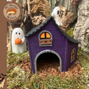 Peut inclure: Une maison miniature violette avec un toit noir, une grande porte arquée et deux fenêtres à lumière orange. Une figurine de fantôme blanc est assise sur le toit, et un autre fantôme blanc tenant une petite citrouille orange se tient à gauche.