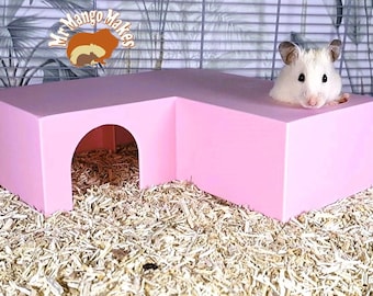 May include: A pink L-shaped hamster hideout with a round entrance. A white hamster is peeking out of the top opening.