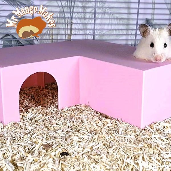 May include: A pink L-shaped hamster hideout with a round entrance. A white hamster is peeking out of the top opening.