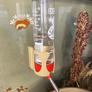 May include: A clear plastic water bottle with a red and silver drinking spout, held in place by a beige plastic holder. The bottle has measurement markings and a label. The image also includes a logo that says "Mr Mango Makes".