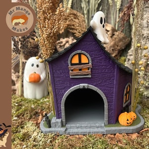 Peut inclure: Une figurine de maison hantée violette avec un toit noir, un fantôme blanc perché sur le dessus et une grande porte arquée. La structure présente des fenêtres éclairées en orange, un volet en bois et deux petites citrouilles orange à la base.