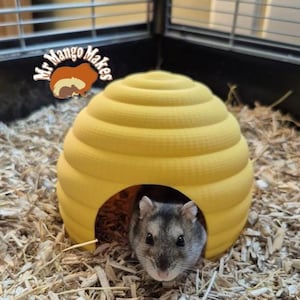 May include: A yellow, beehive-shaped hamster house with a small entrance. A grey and white hamster is visible inside. The house is in a cage with wood shavings. The logo "Mr Mango Makes" is in the top left corner.