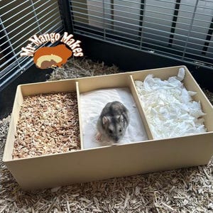 May include: A beige hamster cage with three compartments. One section contains wood chips, another has sand, and the third is filled with white paper bedding. A small grey and white hamster is in the sand section. The cage is inside a larger wire enclosure. The logo "Mr. Mango Makes" is in the top left corner.